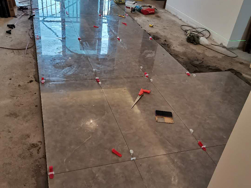 客廳地磚鋪貼：地磚鋪貼前，應(yīng)將地面清掃干凈，尤其是凝結(jié)后的混凝土，否則，地磚鋪貼后會(huì)出現(xiàn)翹邊、空鼓等現(xiàn)象，然后再灑水潤(rùn)濕地面。地面充分濕潤(rùn)，但不能有積水，然后將3:1混合好的沙子水泥鋪在地面上，進(jìn)行基層處理。在地磚背面均勻抹水泥砂漿作為粘合層，厚度不低于1cm，然后鋪在剛才試鋪的位置上，用橡皮錘輕敲地磚飾面直至密實(shí)平整達(dá)到要求。地磚鋪完后24h進(jìn)行清理勾縫，勾縫前應(yīng)先將地磚縫隙內(nèi)雜質(zhì)擦凈，用專用填縫劑勾縫，完工后（一般宜在24h之后）再用棉紗等物對(duì)地磚表面進(jìn)行清理。